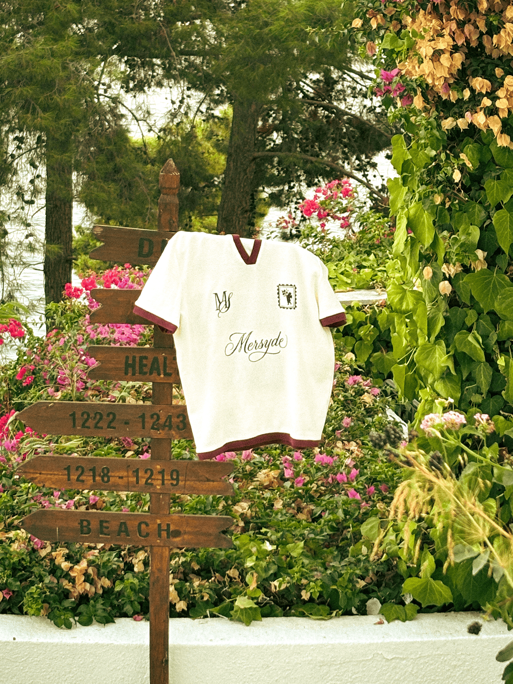 Mersyde cream polo shirt with burgundy trim hanging on a wooden signpost among flowers